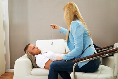 A woman in a blue jacket is performing hypnosis on a man lying on a white sofa, using a swinging pendulum.