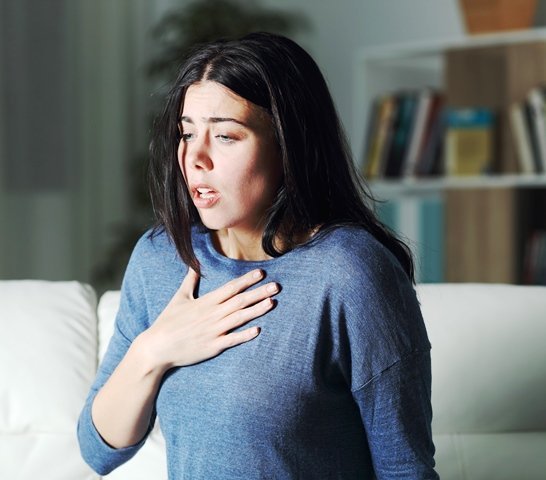 A woman with long dark hair and a blue shirt is sitting on a couch, holding her chest with one hand and looking distressed. A bookcase is visible in the background.