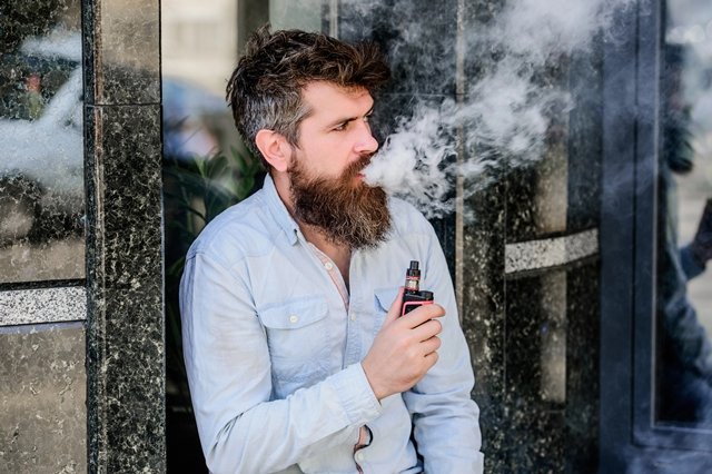 A bearded man in a light shirt sits outside against a marble wall, exhaling a large cloud of vapour from an electronic cigarette.