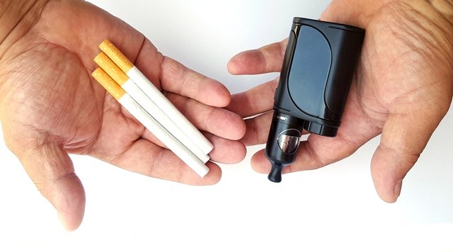 Two hands are shown: the left hand holds three cigarettes, and the right hand holds a black vaping device, both on a white background.