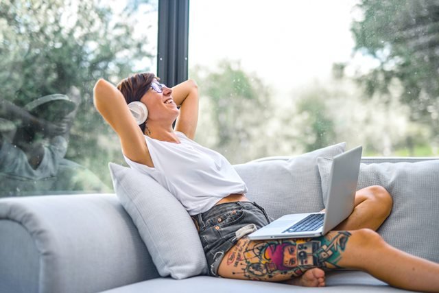 A person with tattoos relaxes on a sofa with a laptop on their lap, wearing headphones and smiling, sitting by a large window with greenery visible outside.
