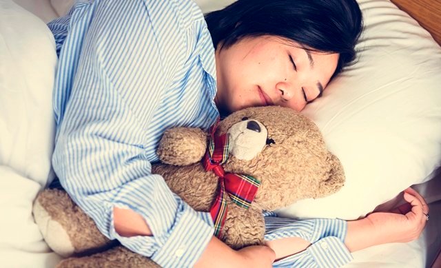 A woman in bed with a teddy bear A person sleeps in bed while cuddling a teddy bear, wearing a striped pajama top.